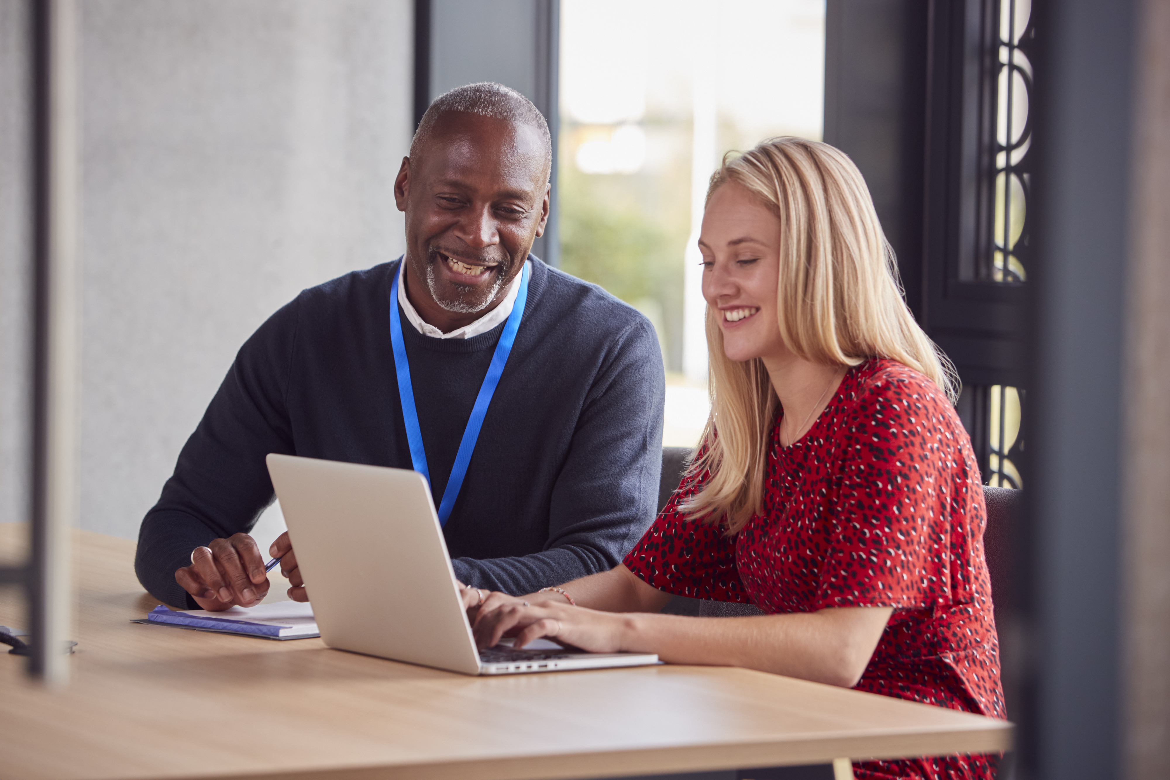 Two people, one wearing a lanyard, smiling and working on a laptop at a desk