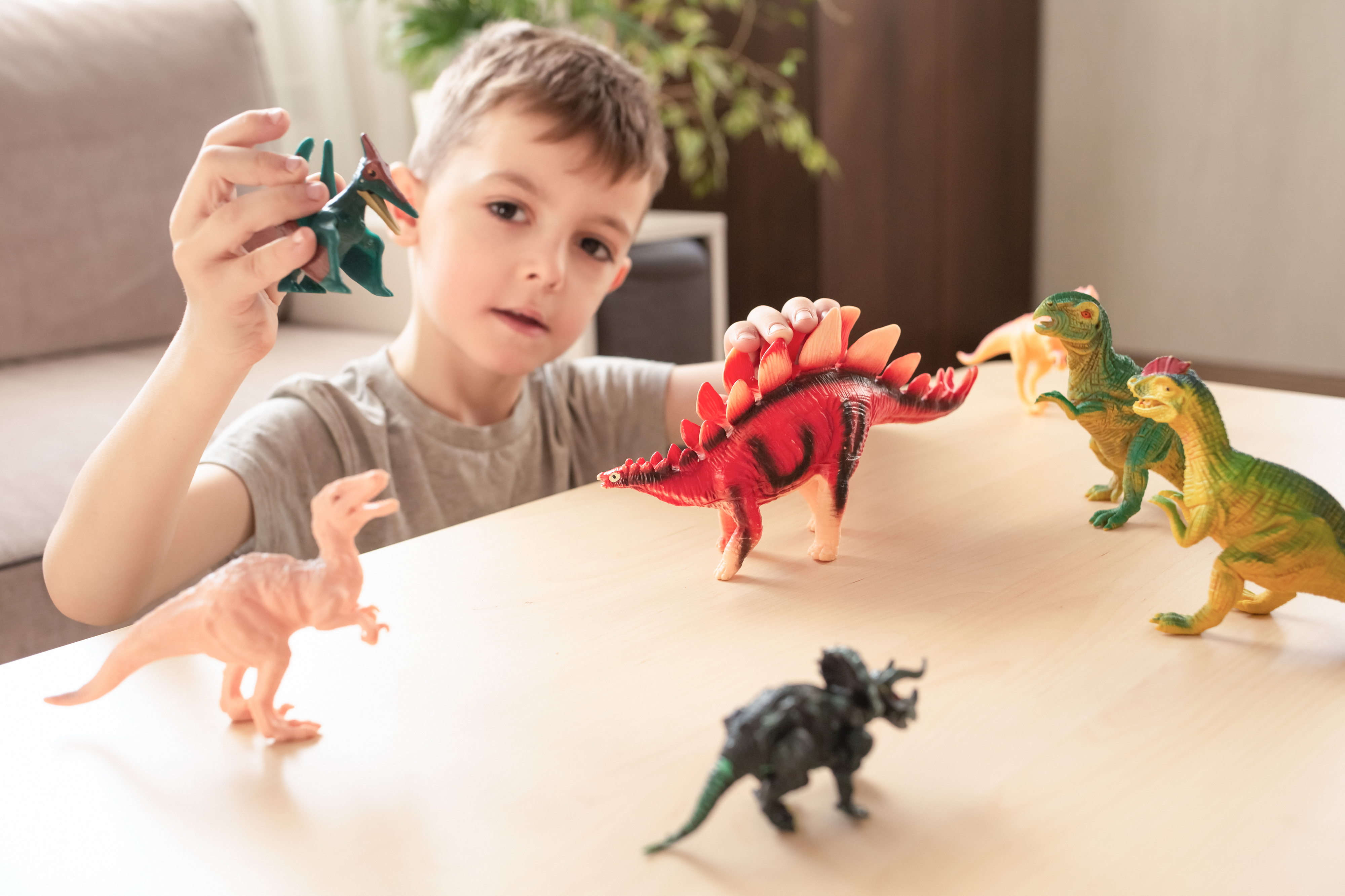 A young boy plays with various dinosaur toys placed on a table, including a Stegosaurus and Pterodactyl, engaging in imaginative play