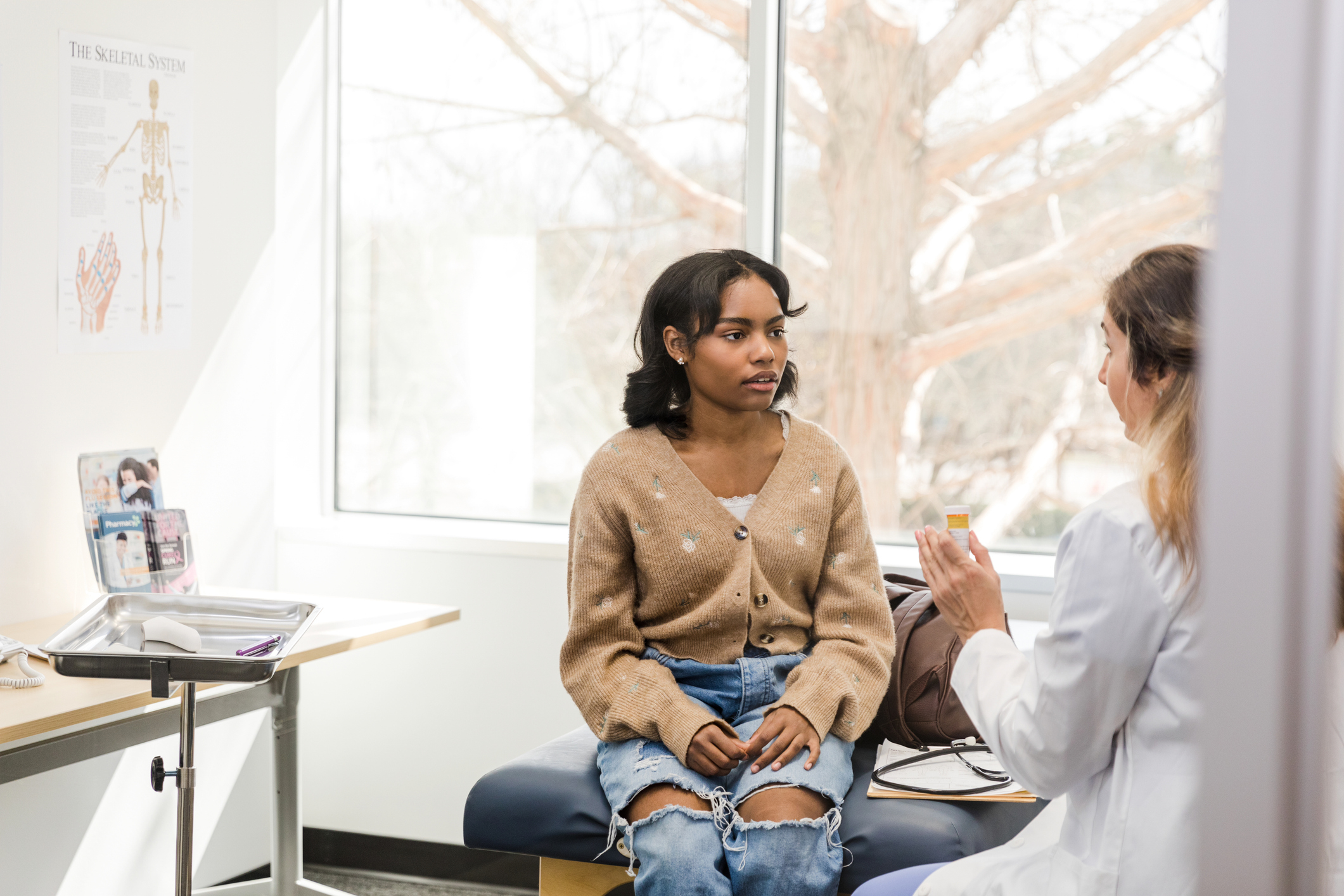A doctor wearing a white coat speaks with a woman in a beige cardigan and ripped jeans sitting on an exam table in a bright medical office