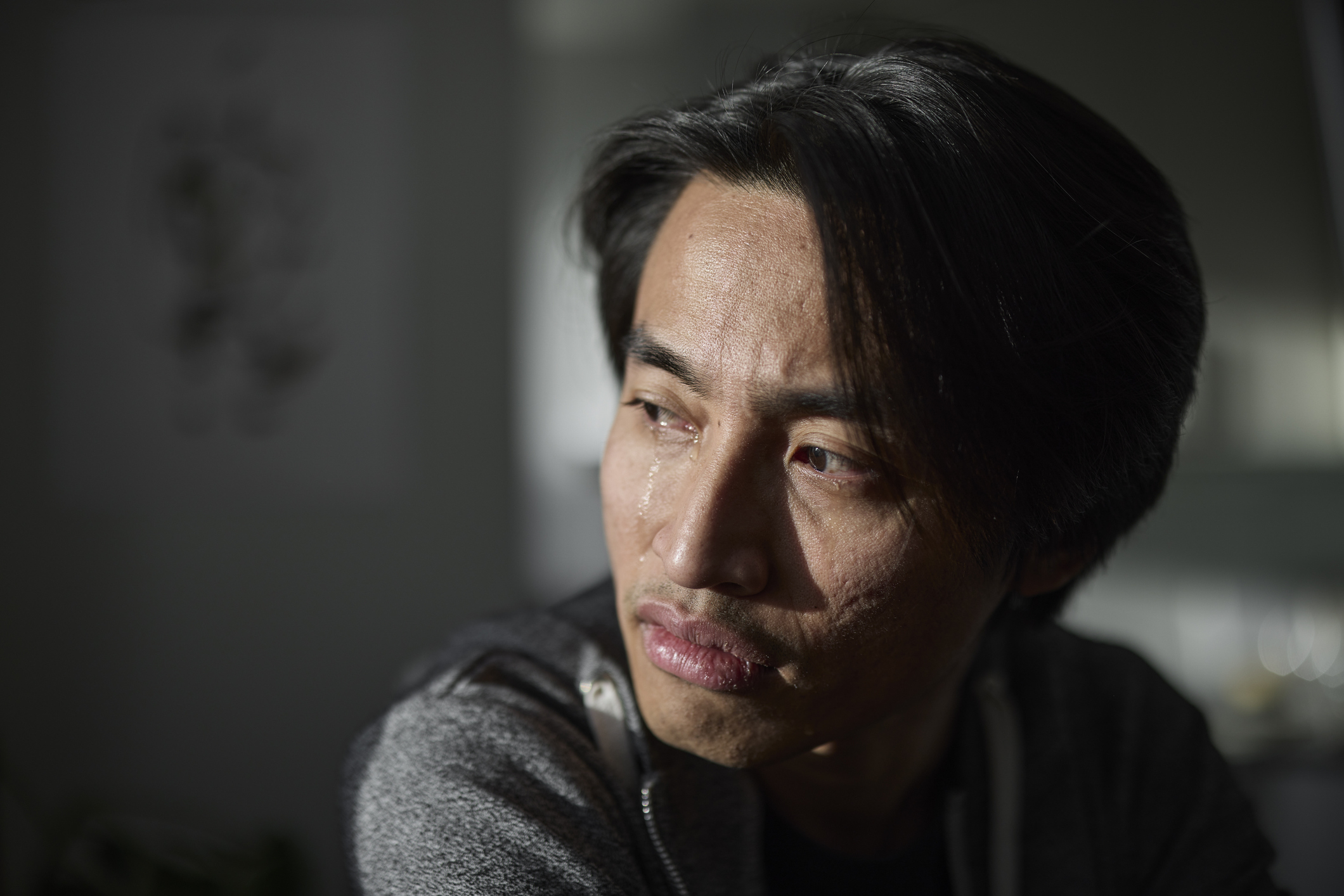 A man with a pensive expression, looking to the side. He has medium-length black hair and is wearing a casual hoodie. The background is blurred