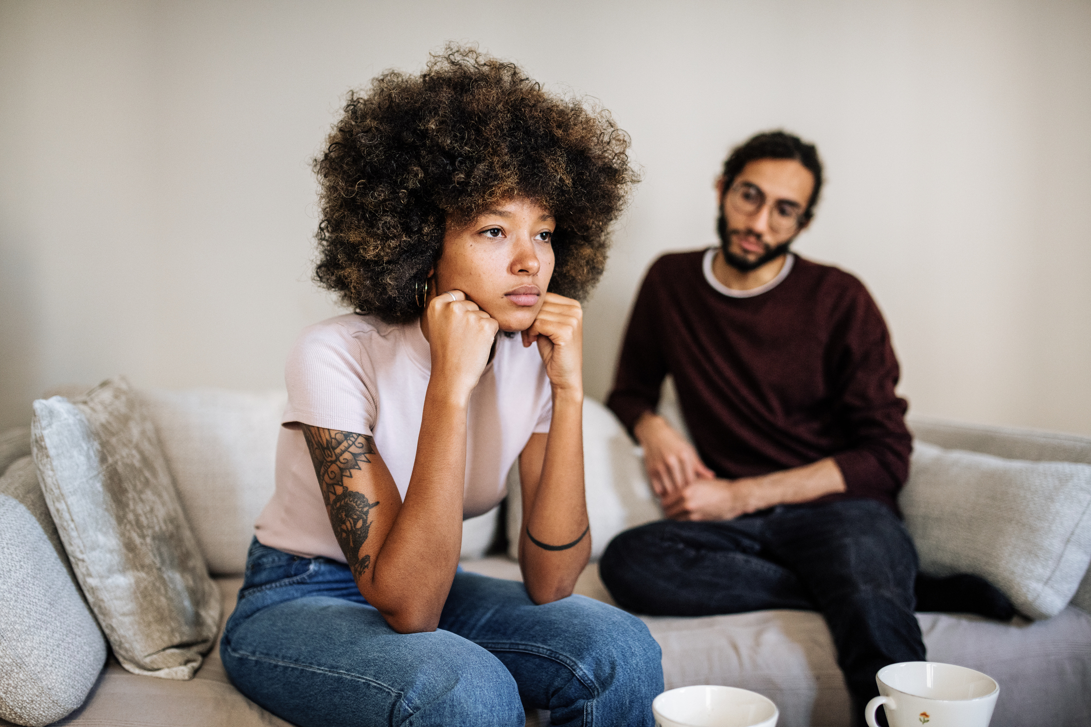 A woman with curly hair and tattoos sits on a couch looking thoughtful, with a man wearing glasses and a sweater sitting behind her, holding his hands together. Cups are on the table in front of them
