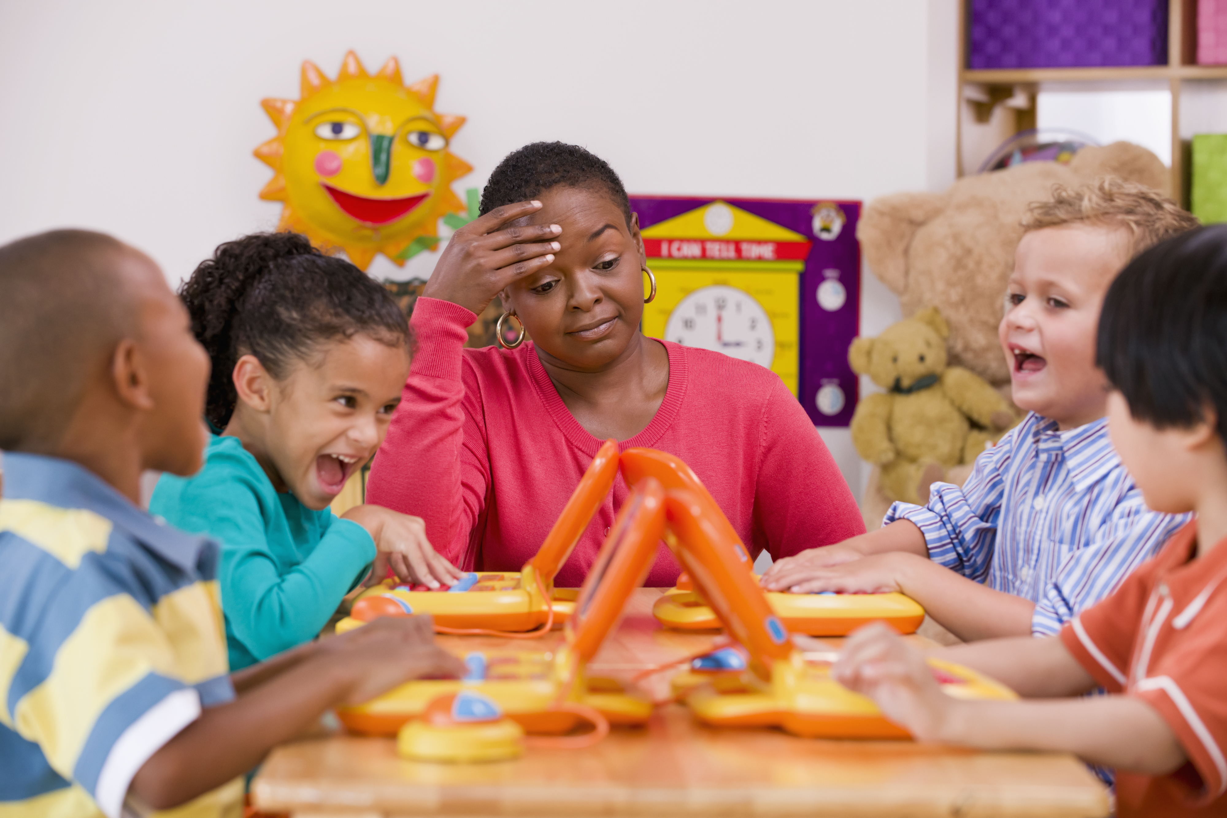 Teacher sits frustrated at table with four children laughing and playing educational games. A sun wall decoration and clock are seen in the background