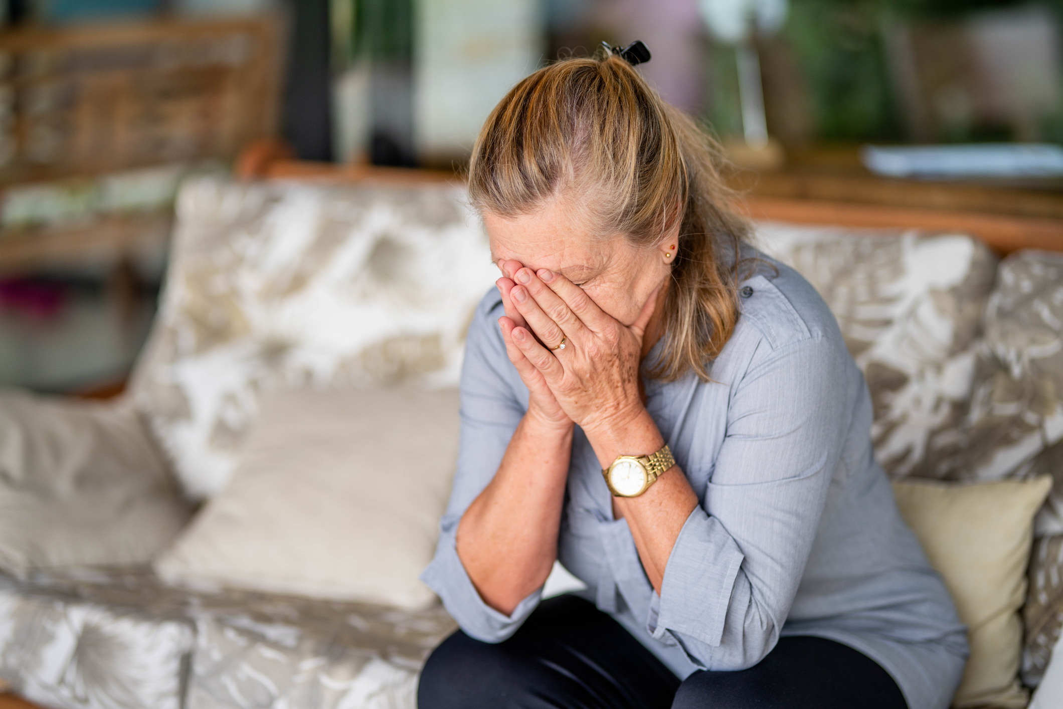 A woman sitting on a couch covers her face with her hands, showing an emotional expression. She is wearing a grey blouse and a watch on her left wrist