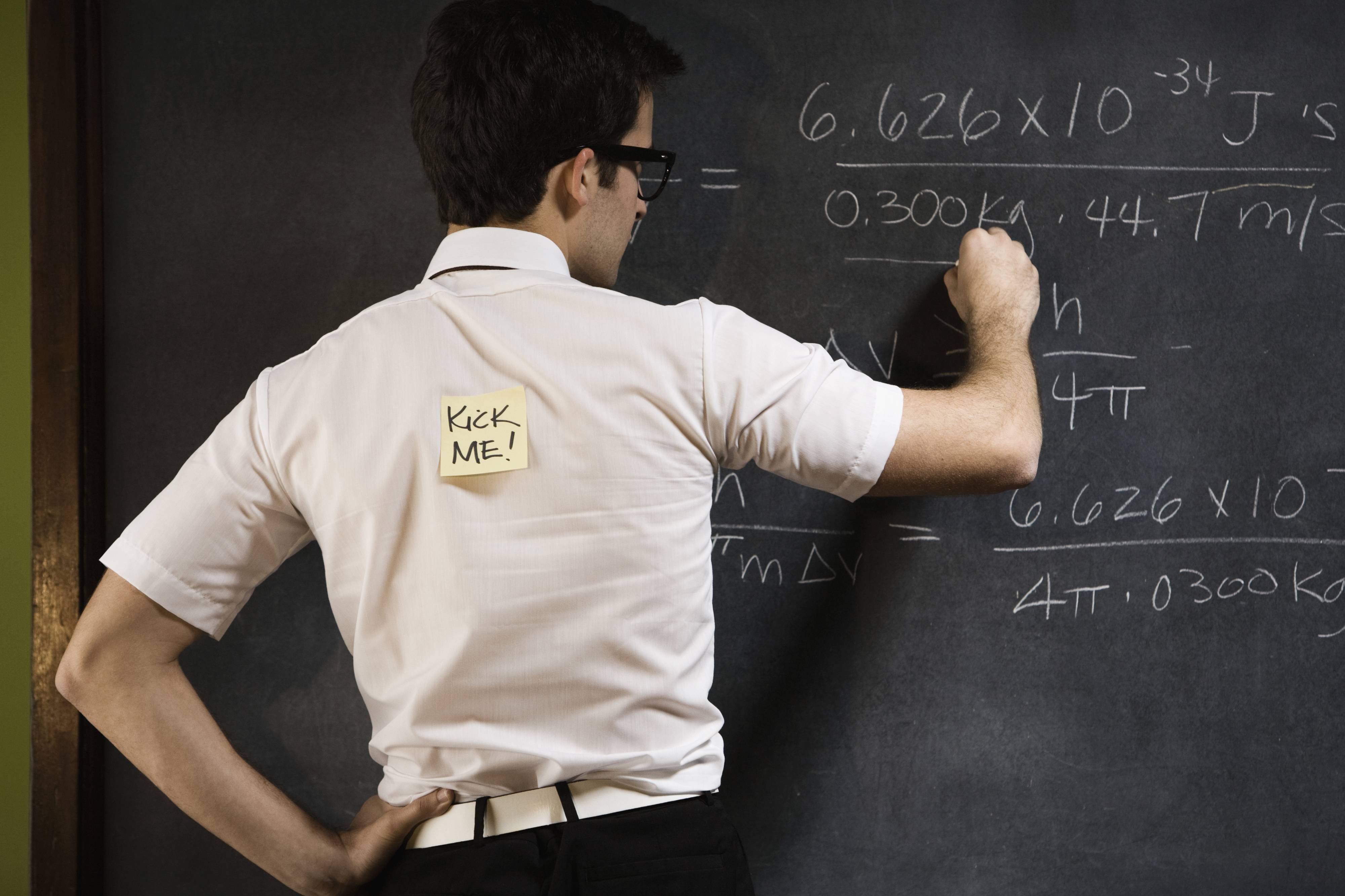 A man with glasses is writing equations on a chalkboard. There is a sticky note on his back that reads "Kick Me!"