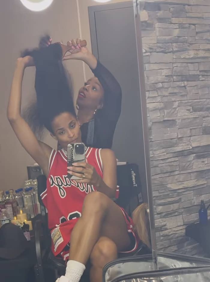 Ciara is sitting in a chair wearing a sports jersey, holding her phone up to a mirror. A hairstylist stands behind her, working on her hair