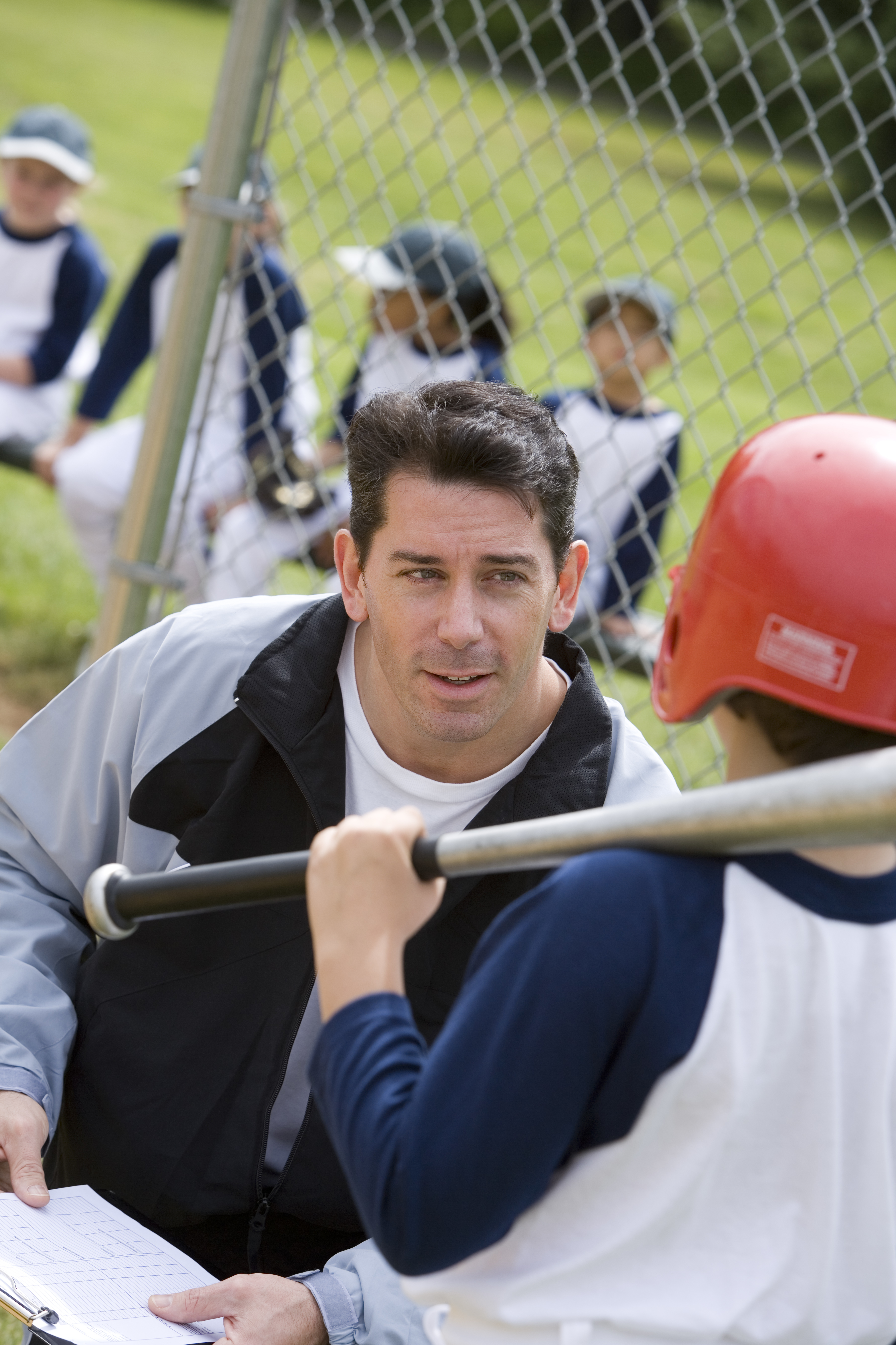 An adult in a jacket talks to a young baseball player in a helmet, holding a bat. Other children in baseball uniforms sit in the background near a fence. Names unknown