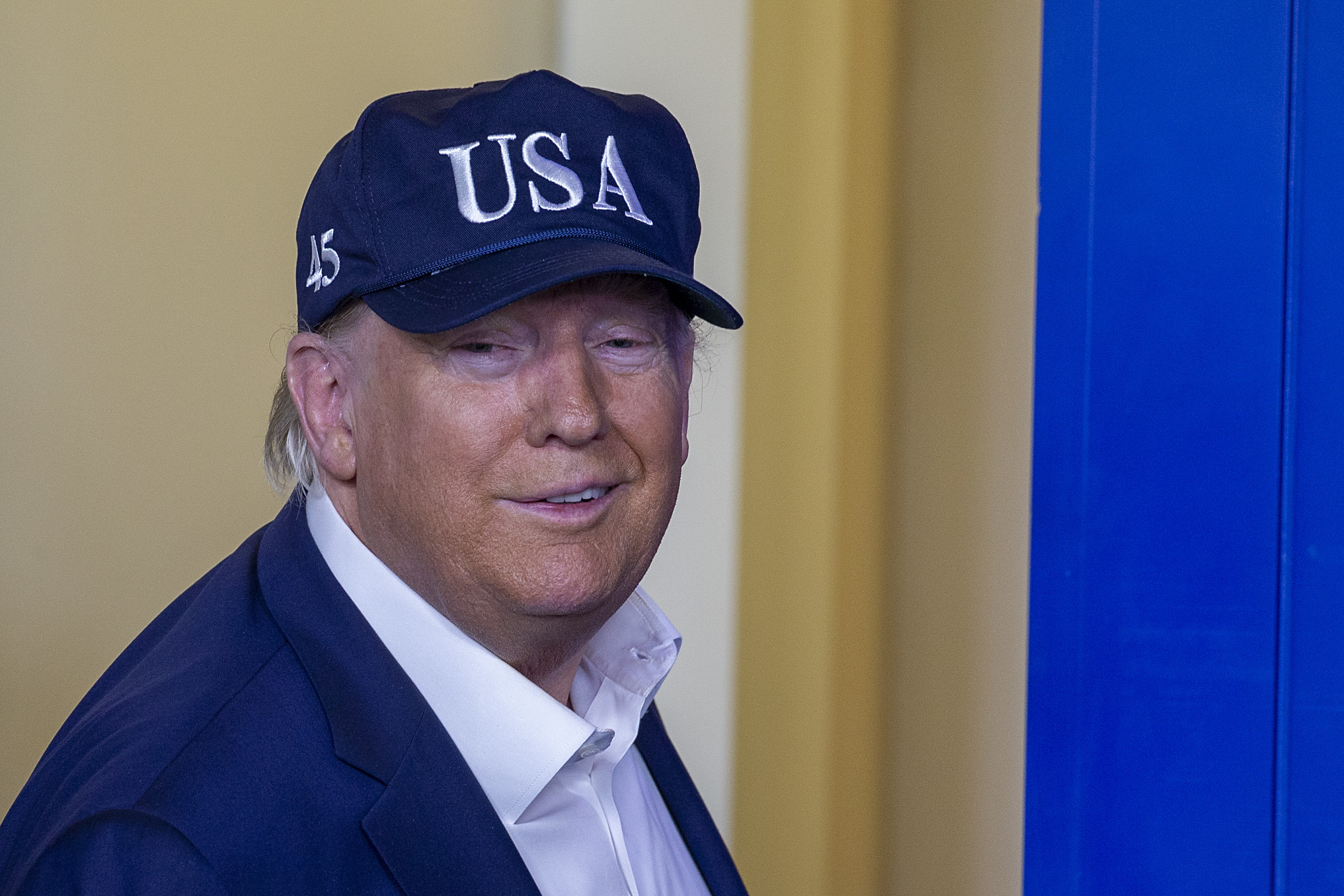 Donald Trump wearing a blue jacket and a black "USA" baseball cap with "45" on the side, smiling