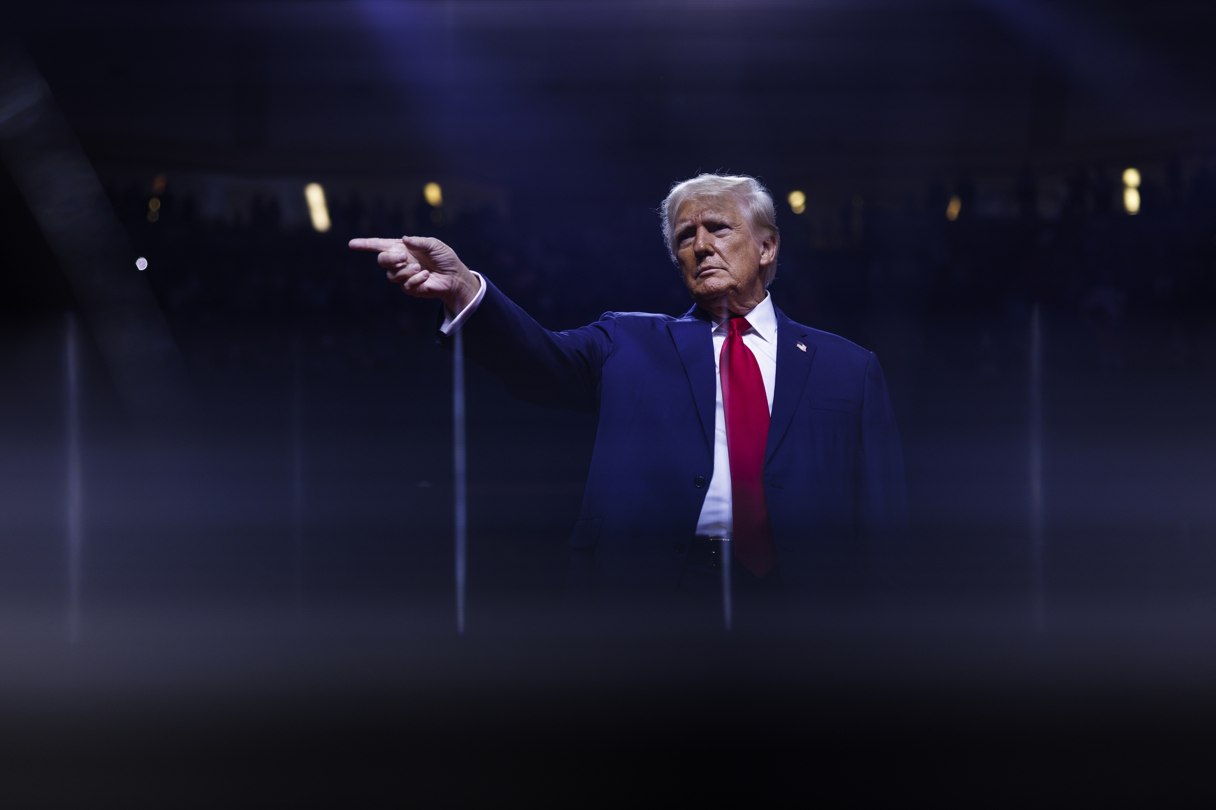 Donald Trump is pointing with his right hand while standing under stage lights. He is wearing a suit with a red tie, and the background is dark and hazy