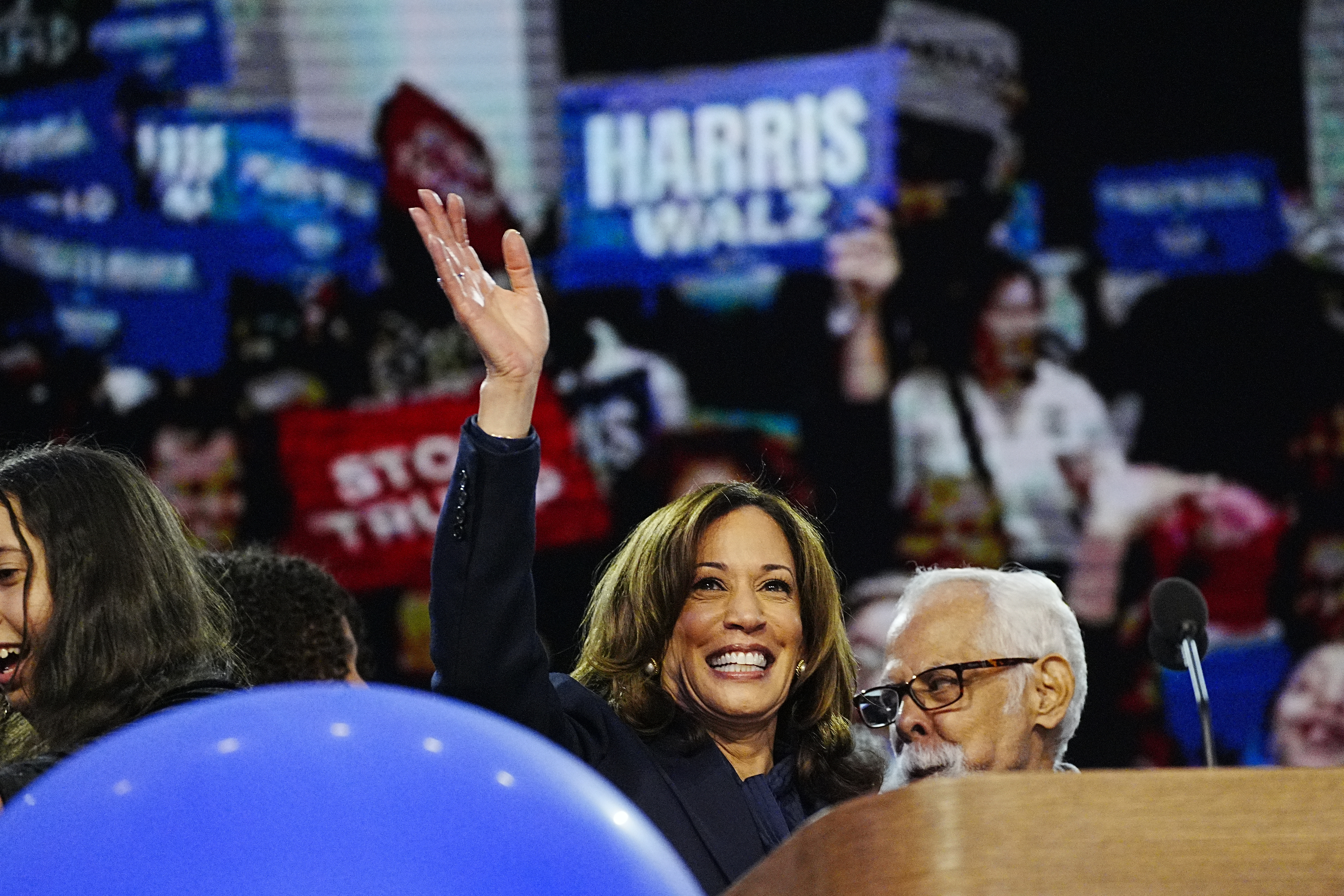 Kamala Harris waves to a crowd holding “Harris” signs at a rally, accompanied by another individual at a podium
