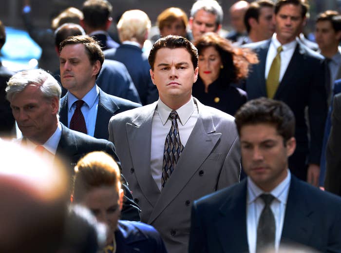 Leonardo DiCaprio, in a business suit, walks confidently in a crowded street scene, featuring numerous people in professional attire