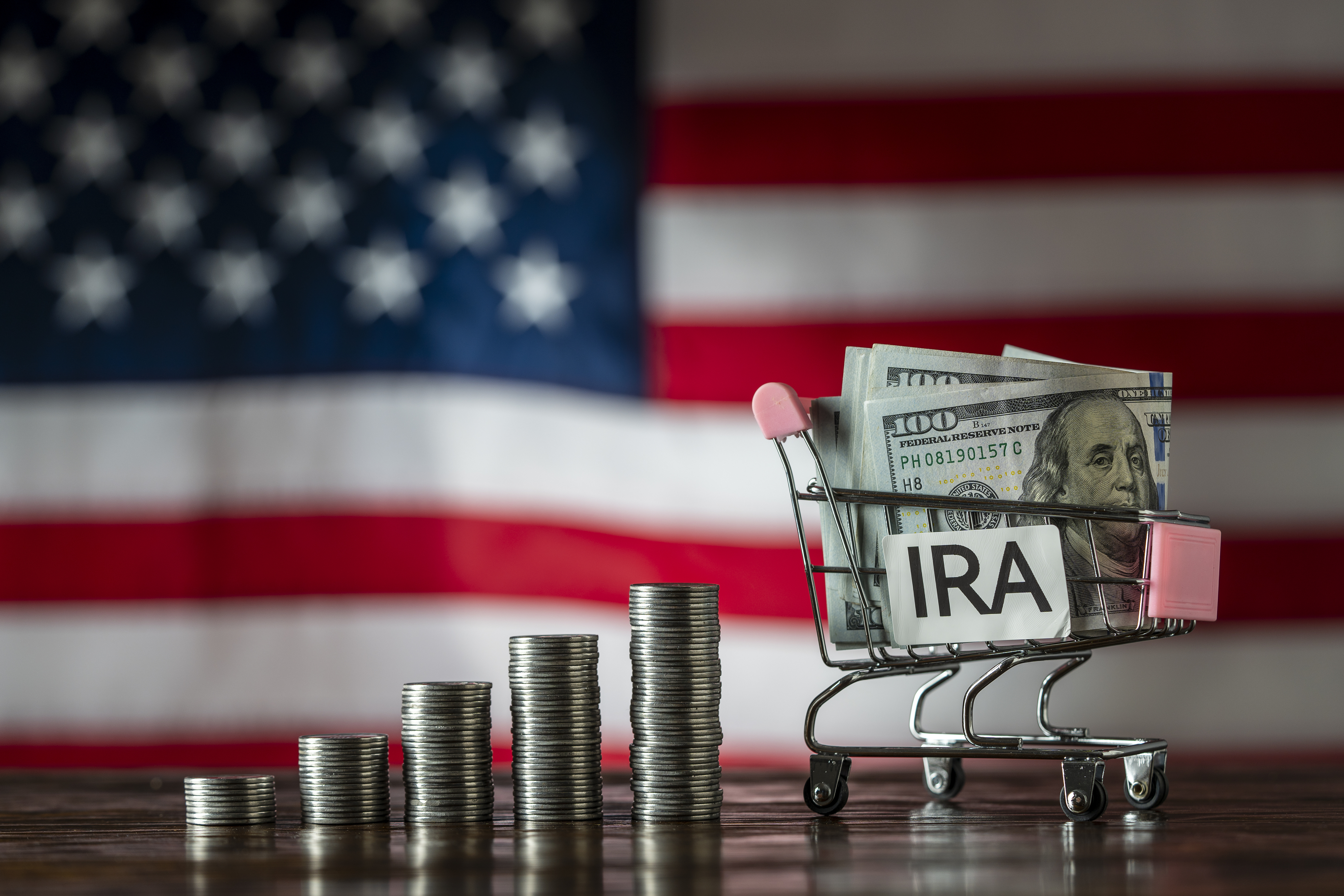 A small shopping cart labeled "IRA" is filled with $100 bills. Stacks of coins increase in height next to it, with an American flag in the background