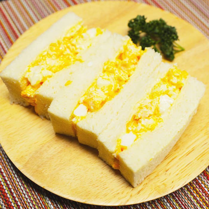 白い食パンに卵サラダが挟まれたサンドイッチが木の皿に盛り付けられ、パセリが添えられています。