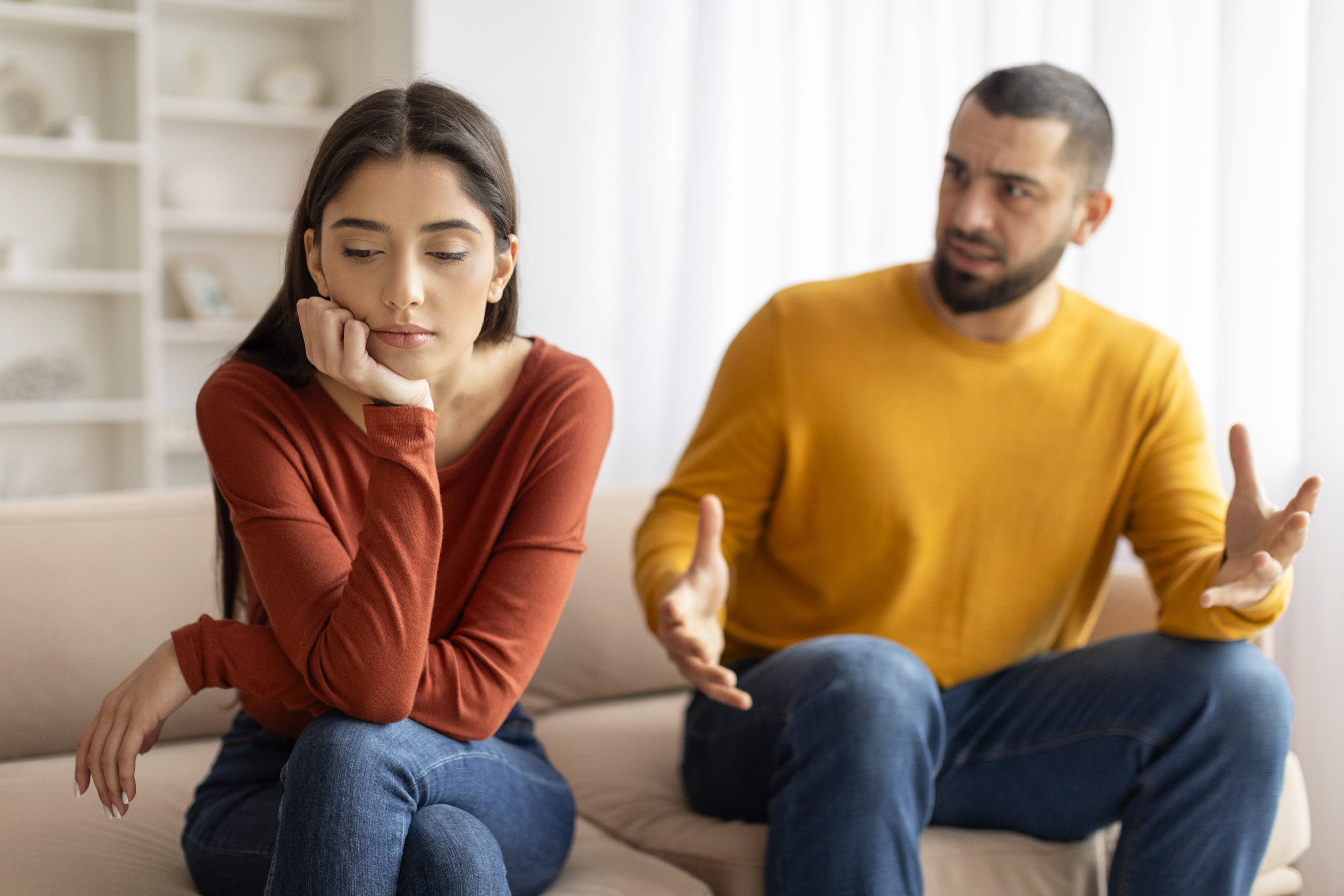 A woman and a man sit on a couch, engaged in a serious conversation. The woman looks away with her hand on her cheek, while the man gestures openly