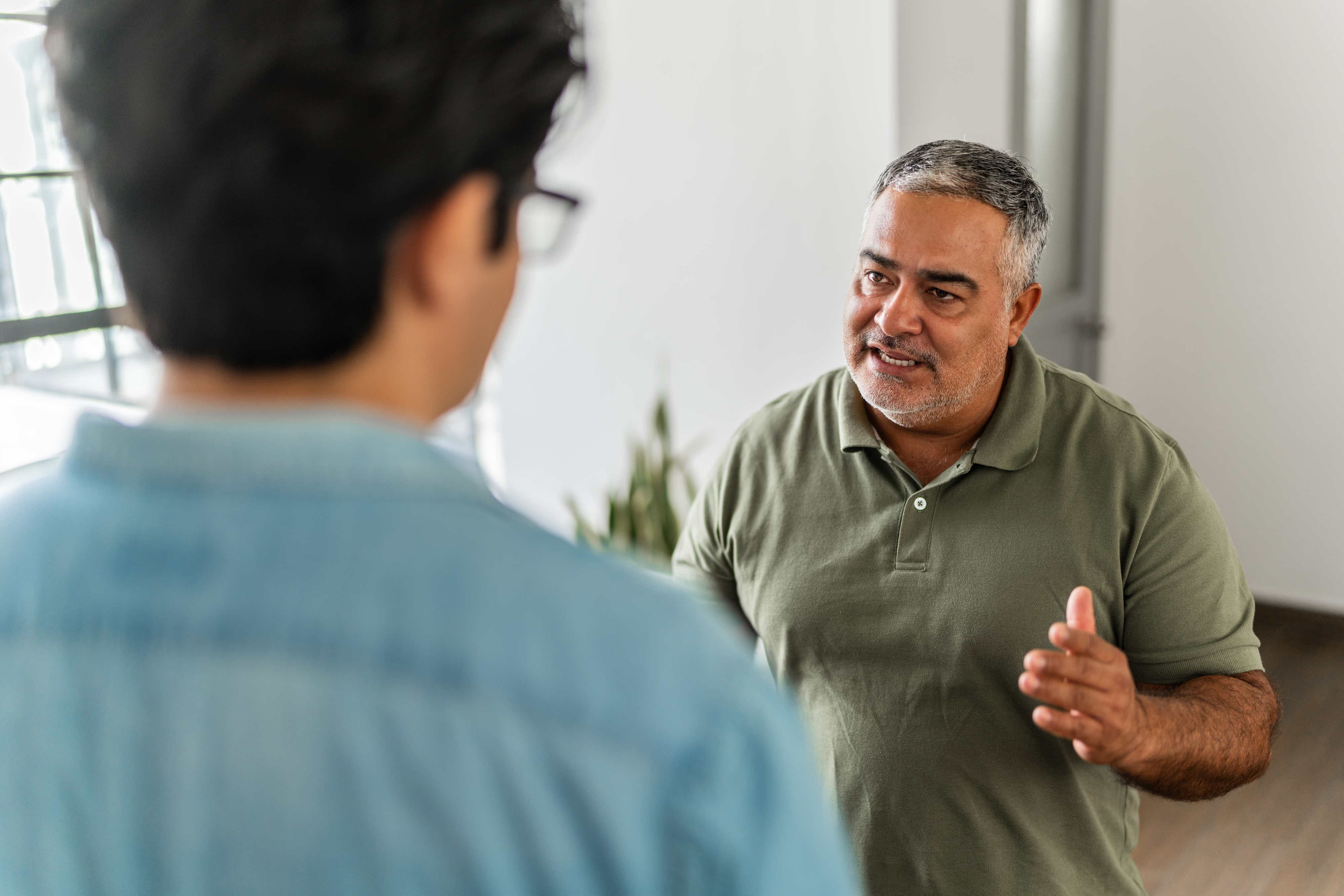 Two people are in conversation at home. One person in the foreground has their back to the camera, and the other, in a polo shirt, is talking animatedly