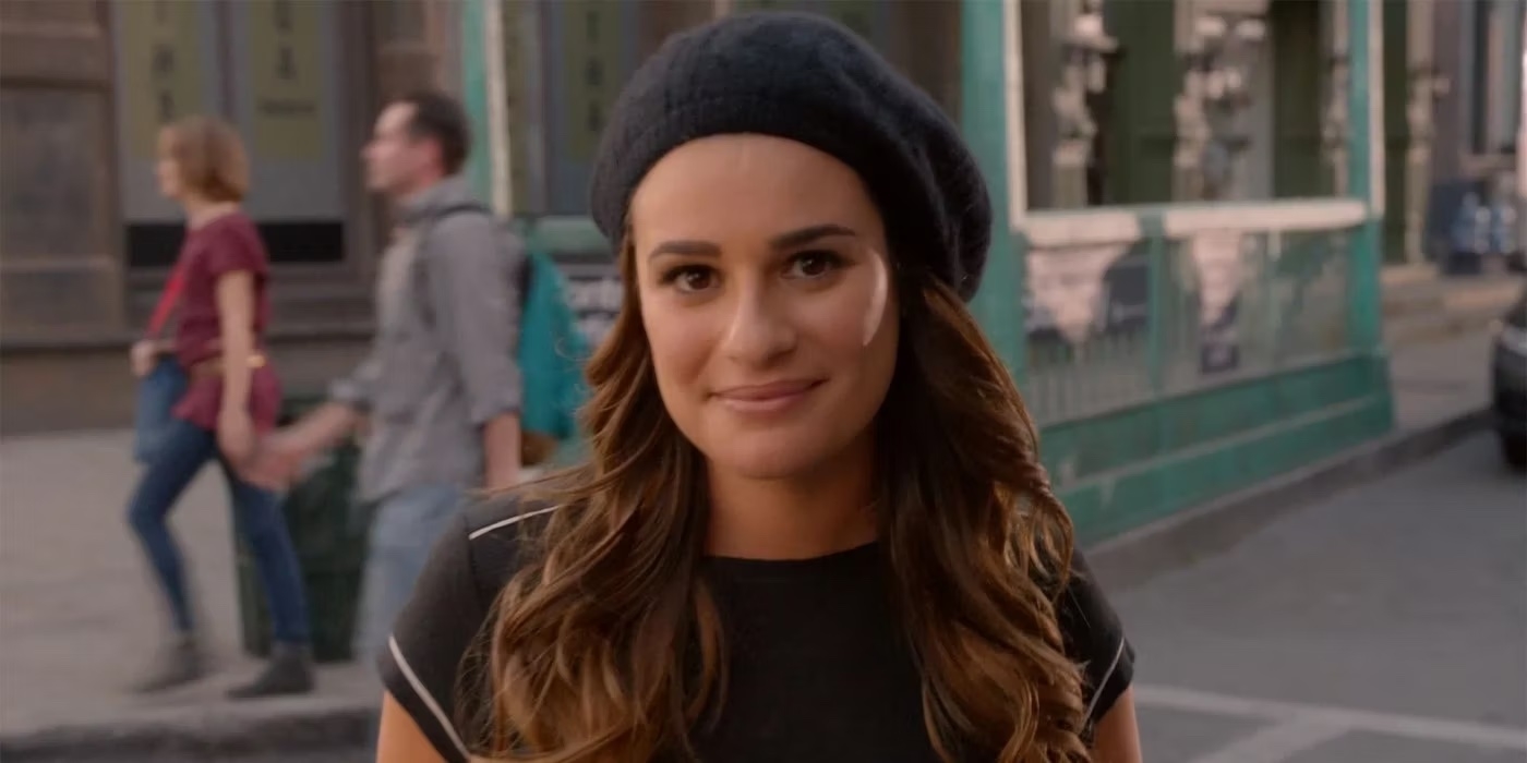 Lea Michele wearing a black beret and a casual top, smiling and outdoors with blurred pedestrians in the background