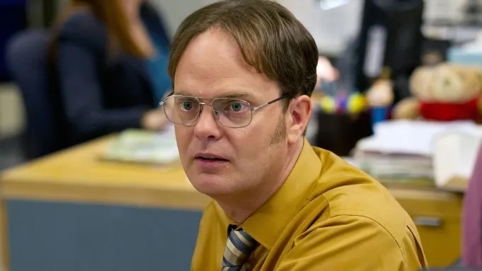 Rainn Wilson as Dwight Schrute in an office setting from the TV show "The Office," wearing a mustard-yellow shirt, striped tie, and glasses