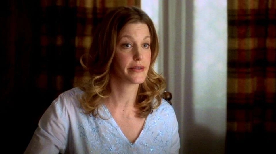 Anna Gunn in a scene from a TV show, wearing a light blouse, seated indoors with patterned curtains in the background
