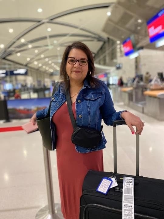 A reviewer wearing the reddish  casual formal  and denim overgarment   stands successful  an airdrome  with a rolling suitcase and a tablet, acceptable   for travel