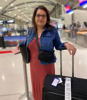 A reviewer wearing the reddish casual formal and denim overgarment stands successful an airdrome with a rolling suitcase and a tablet, acceptable for travel