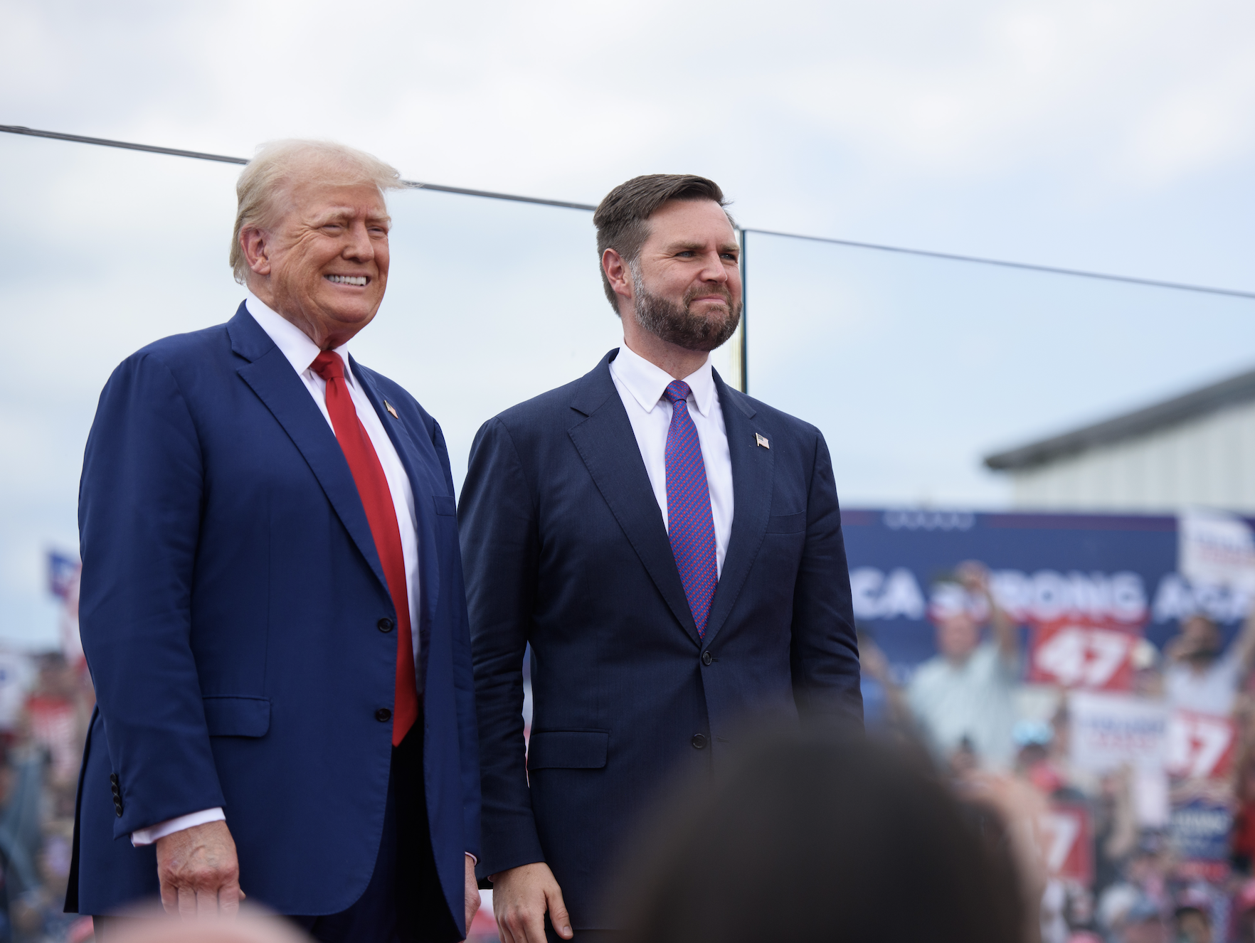 Donald Trump and another man standing together at an outdoor event, both in suits and ties, with a crowd and signage in the background