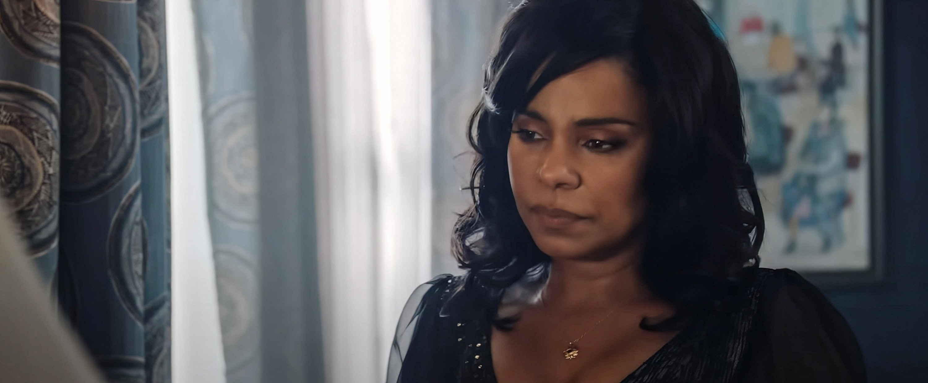 Barbara Jean  wearing a sheer black top with decorative studs, looks thoughtfully to the side in an indoor setting with curtains and a painting visible in the background