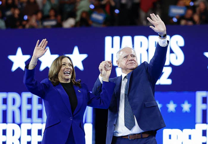 Kamala Harris and Tim Walz wave, holding hands, at a political rally with audience and signs in the background