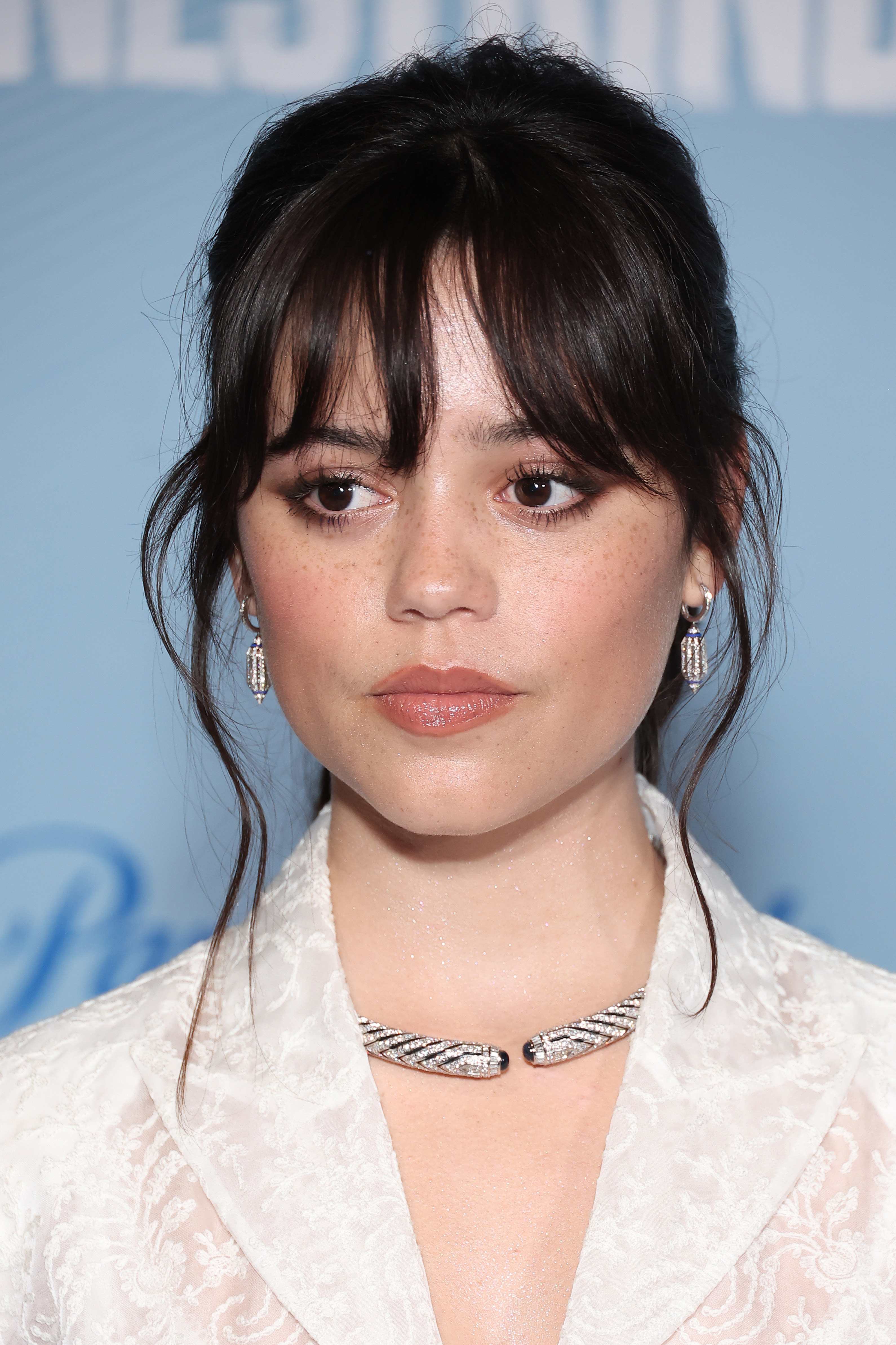 Jenna Ortega at a public event, wearing a lace outfit with a jeweled necklace and earrings