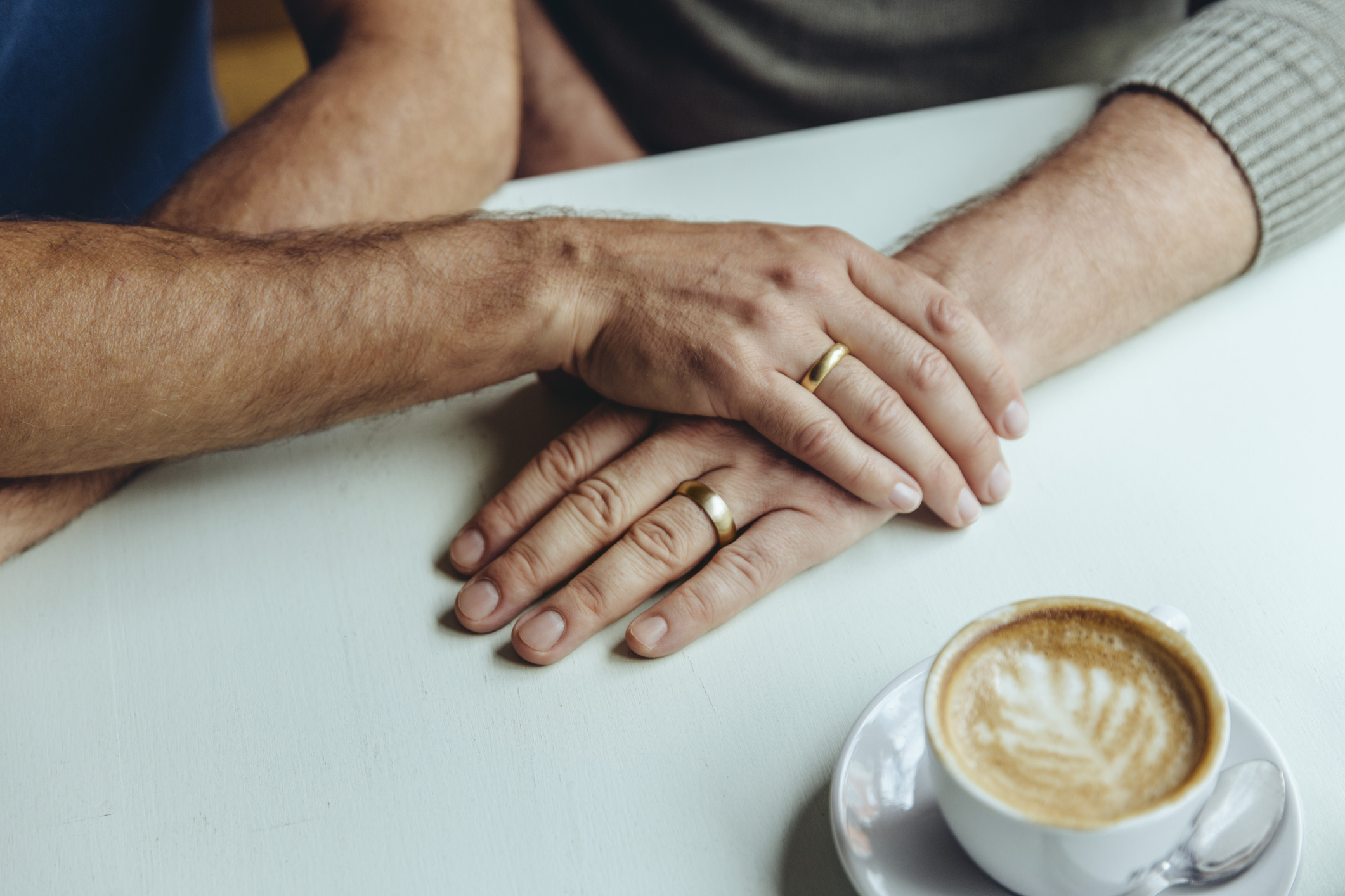 Two hands with wedding rings gently rest on each other, next to a cup of coffee with latte art