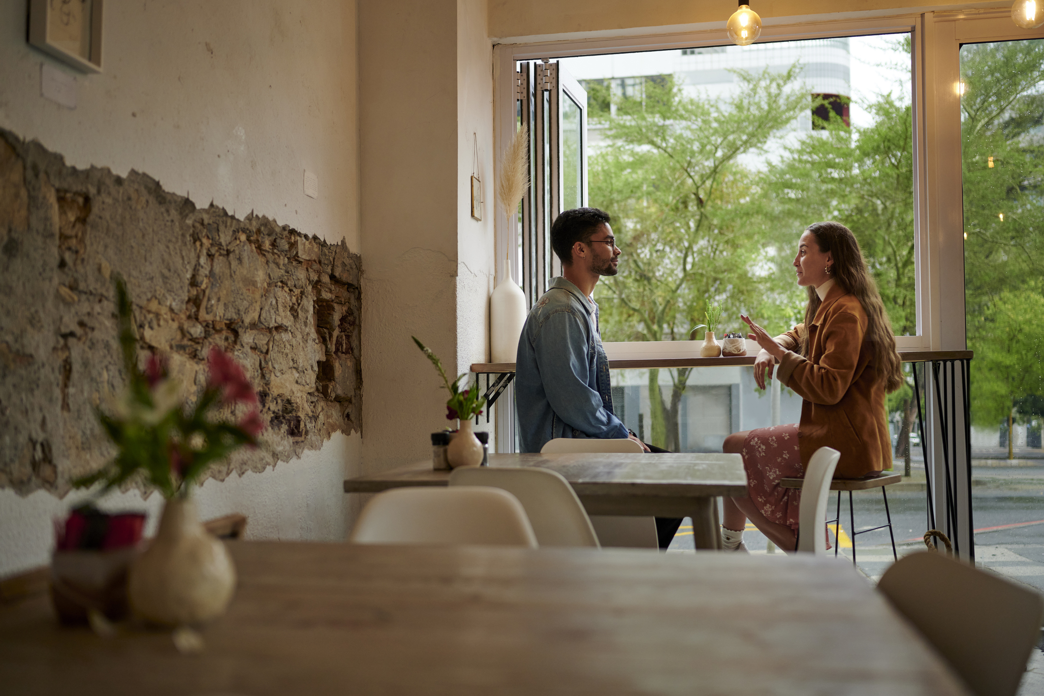 A couple sits at a cozy café, engaged in a lively conversation by the window. The café has a rustic interior with exposed brick walls and vase decorations