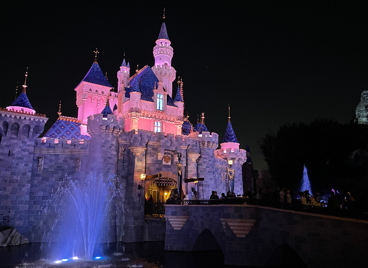 Disneyland castle illuminated at night with light purple and pink hues, with fountains in the foreground and visitors in the distance