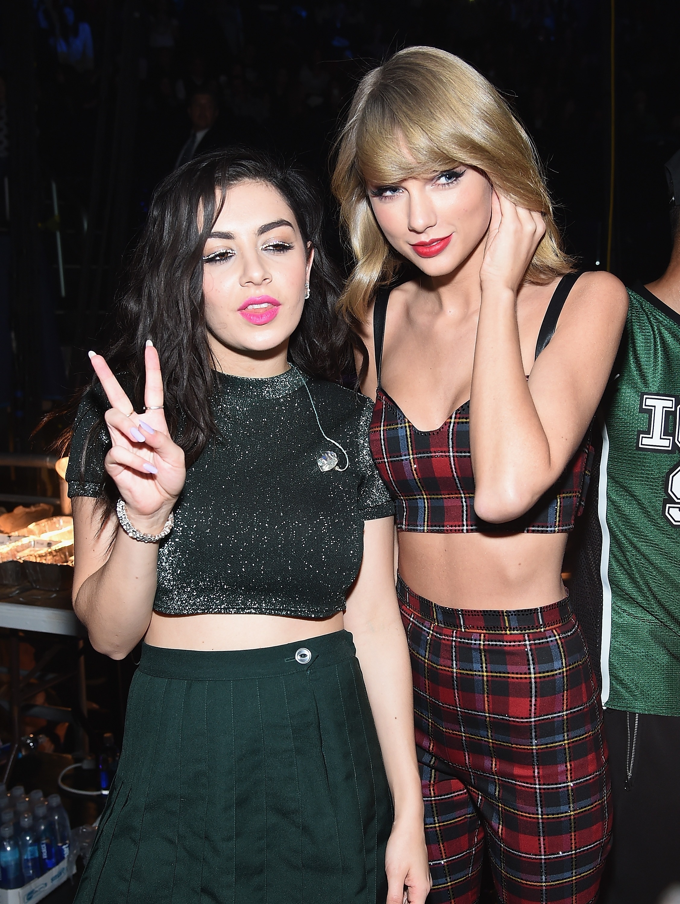 Charli XCX in a sparkly crop top and skirt poses with Taylor Swift in a plaid crop top and pants at an event. Charli makes a peace sign