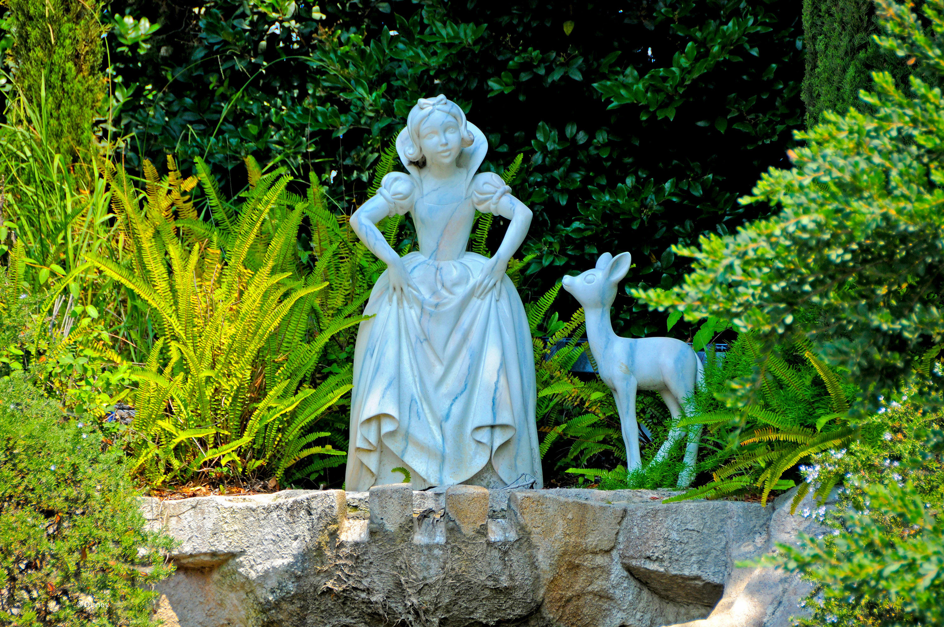 Snow White statue with hands on hips standing beside a deer statue in a lush garden setting with greenery and ferns