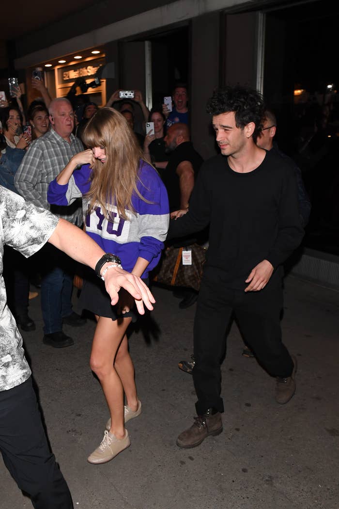 Taylor Swift and Matty Healy hold hands while walking through a crowd. Taylor wears a casual sweater and skirt, and Joe is dressed in a plain, dark outfit