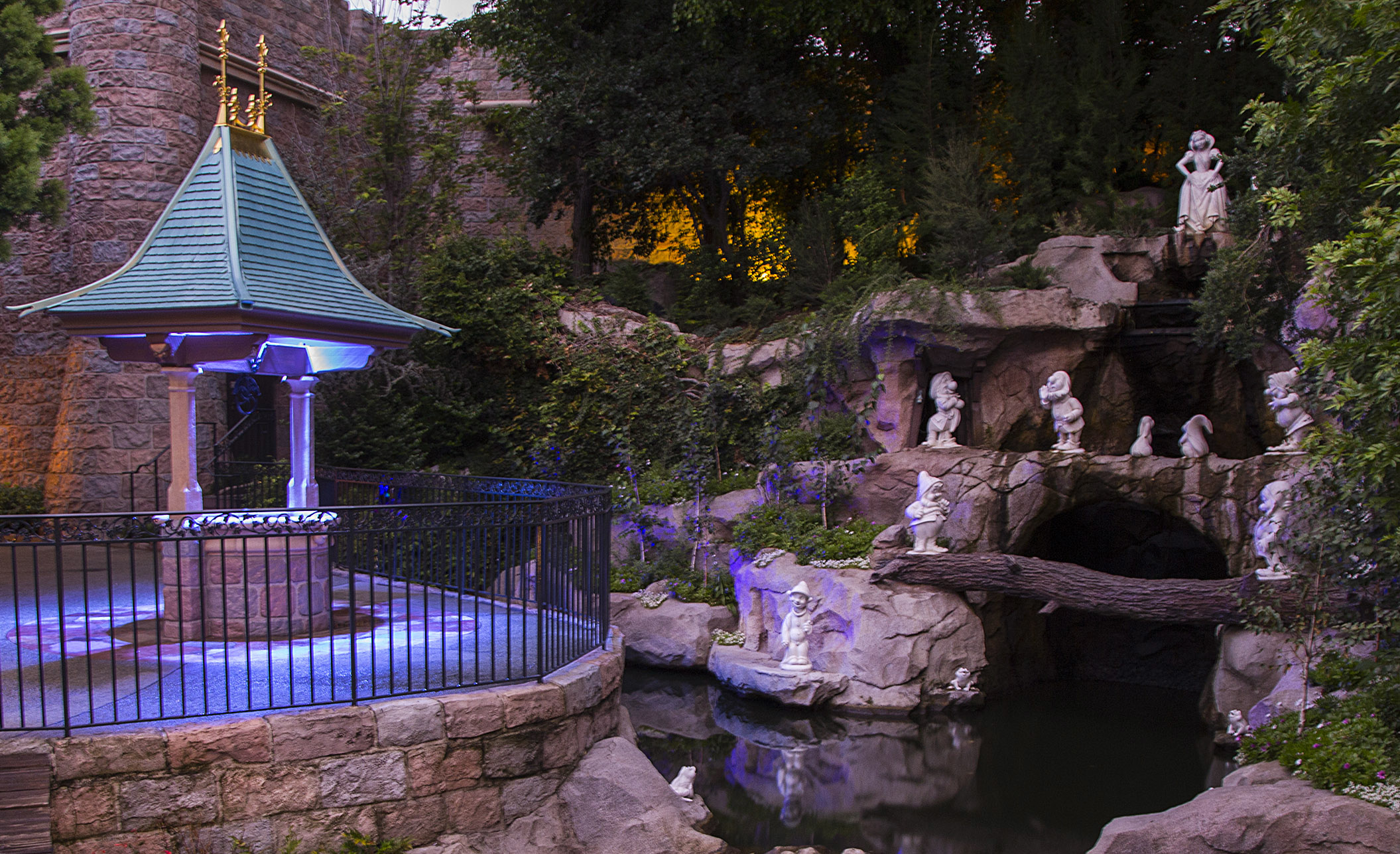 An enchanting garden scene featuring the Snow White Grotto at Disneyland with statues of Snow White and the Seven Dwarfs near a small pond and gazebo