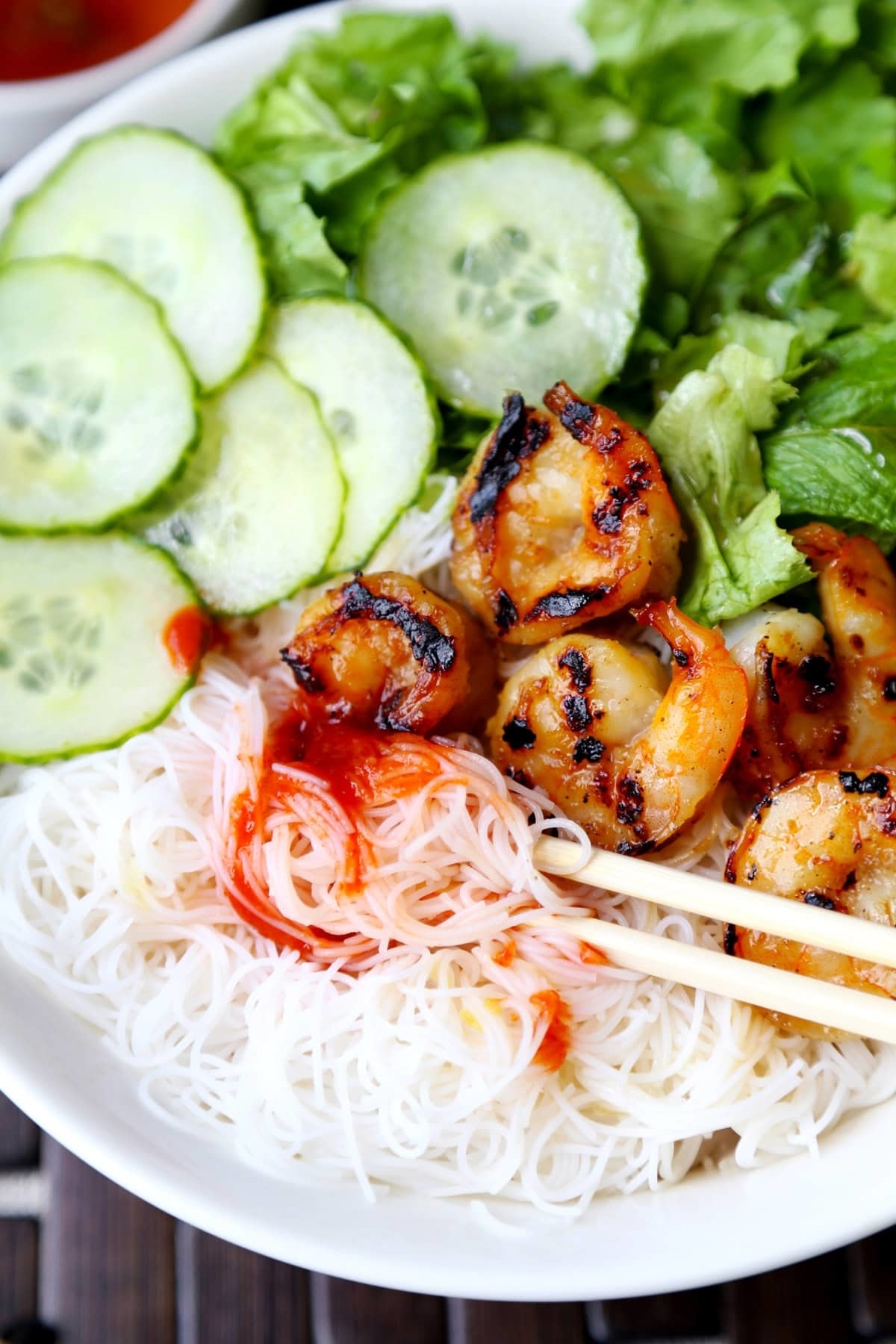 A bowl of vermicelli noodles topped with grilled shrimp, sliced cucumbers, and lettuce, garnished with red sauce, with chopsticks resting on the dish