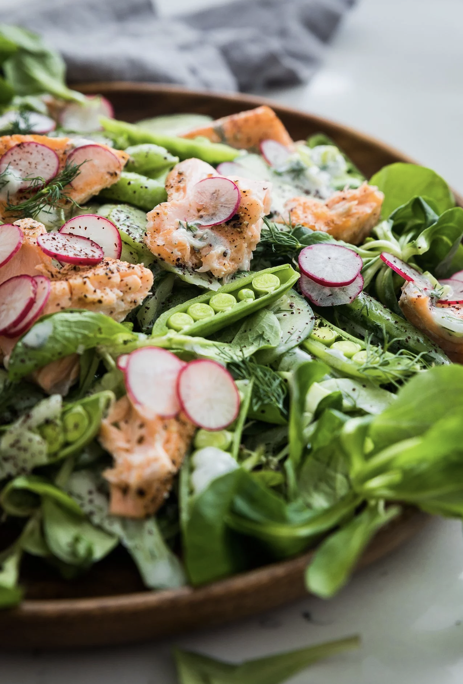 A fresh salad with mixed greens, sliced radishes, snap peas, and grilled salmon pieces, garnished with dill and a light dressing