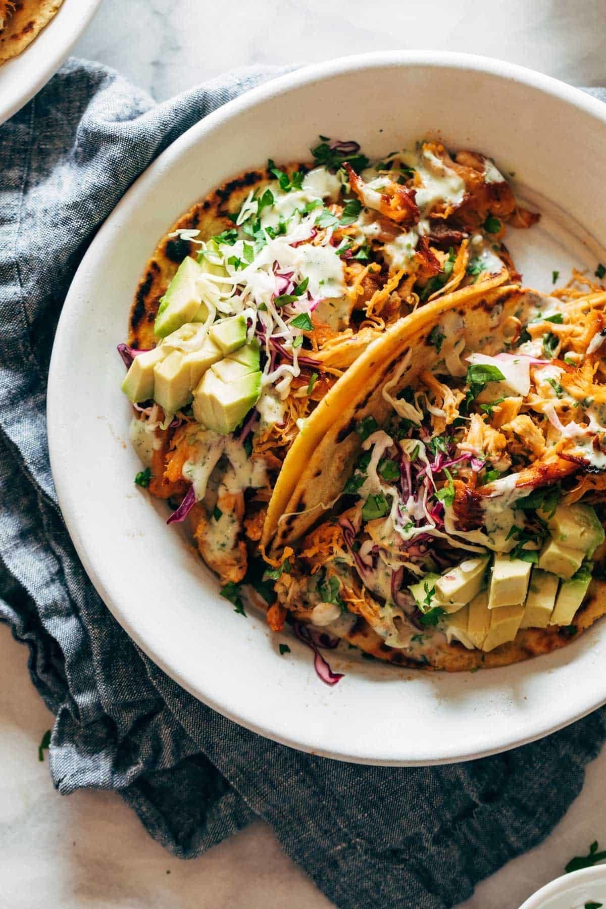 Two tacos on a white plate filled with shredded chicken, chopped avocado, cabbage, and drizzled with creamy sauce, with a gray cloth underneath