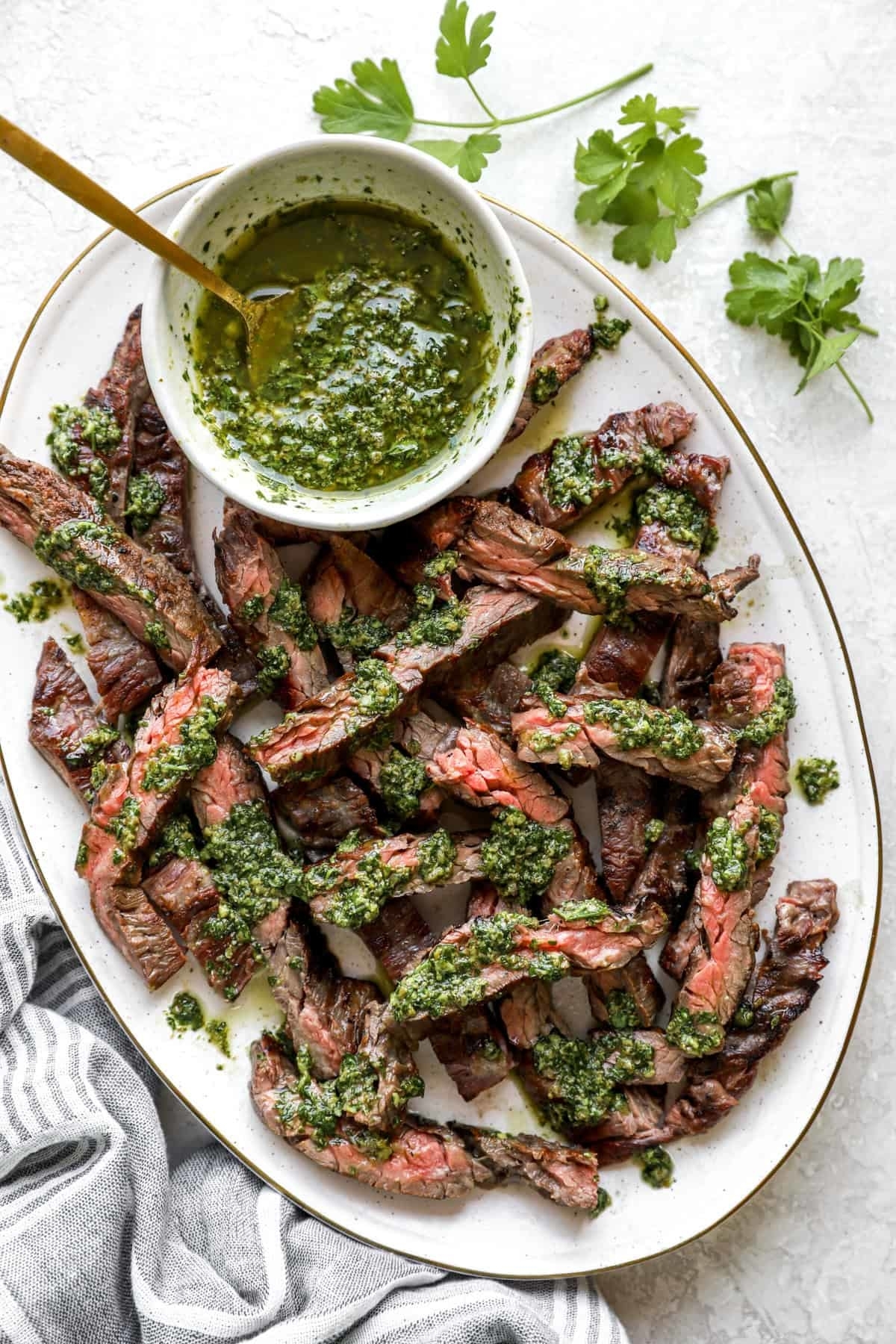 Grilled steak strips drizzled with green chimichurri sauce served on a white plate with a bowl of extra chimichurri on the side. A cloth napkin sits to the left