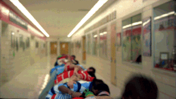 Line of people lying down, wearing sleeping bags, moving together on a school hallway floor
