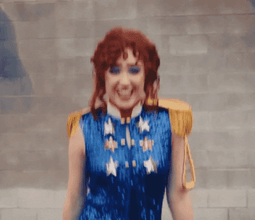 A person with curly red hair wearing a sparkling blue costume with star decorations and gold epaulettes is smiling and dancing excitedly