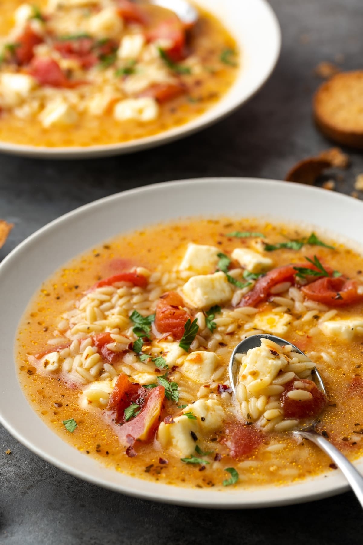 A close-up of two bowls of tomato orzo soup with chunks of cheese, herbs, and a spoon in one bowl. Toasted bread slices are on the side