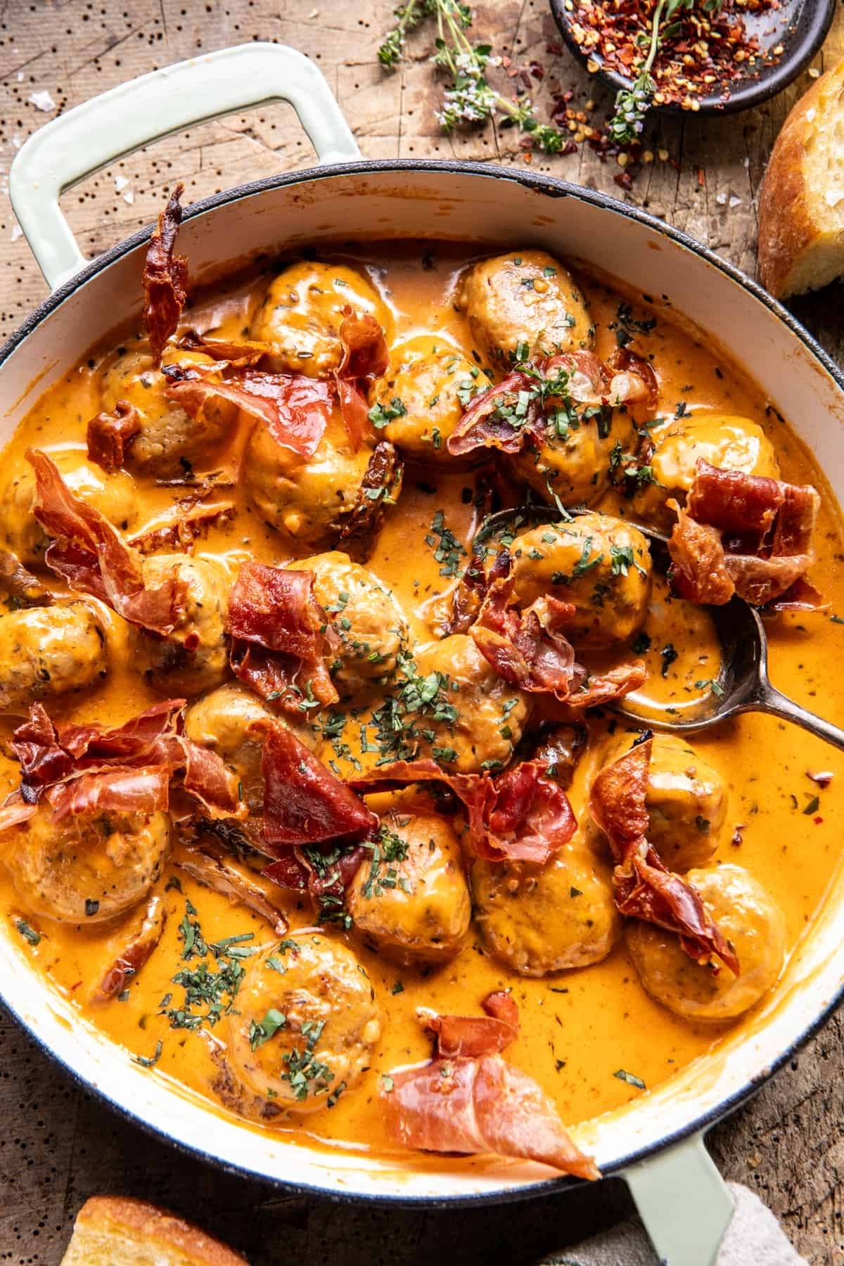 A skillet filled with creamy meatballs garnished with crispy prosciutto and herbs, with a spoon on the side
