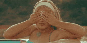 Sabrina Carpenter rides in a car, wearing a heart-shaped necklace and a blue top, playfully covering her eyes with her hands