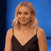 Kristen Bell smiling and shrugging on a blue background. She is wearing a sleeveless top