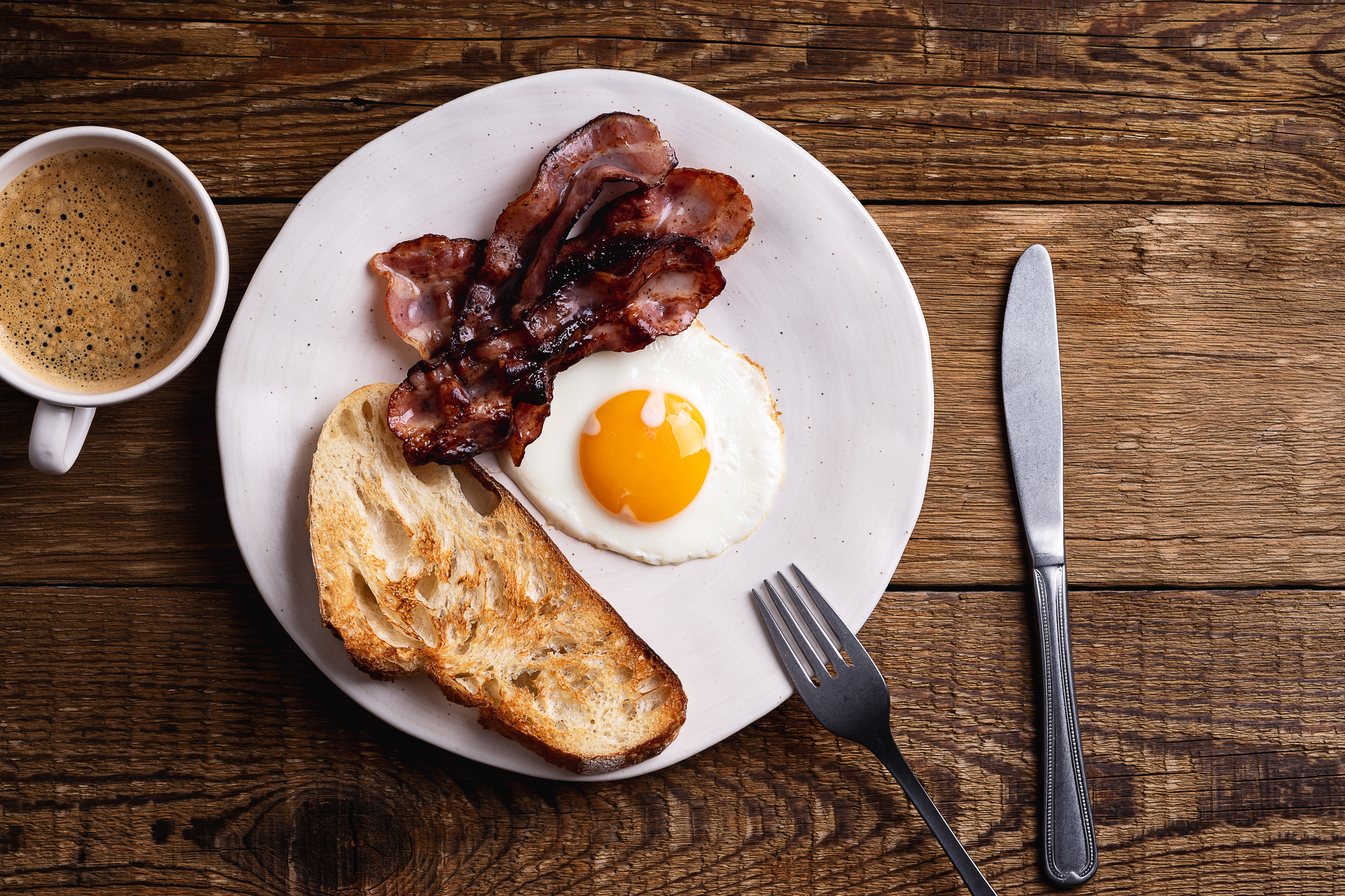 A plate with a fried egg, bacon, and toast beside a cup of coffee on a wooden table