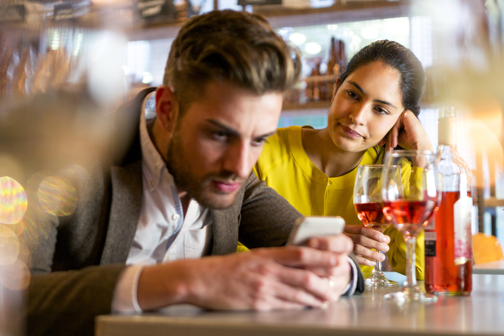 Man looks at phone while woman looks at him, both seated at a bar with wine glasses and bottles in the foreground