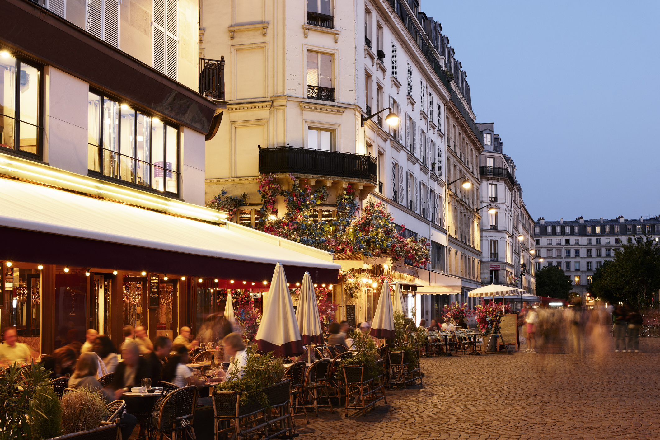 A bustling café-lined street in a European city in the evening, with people dining outdoors and walking along the sidewalk