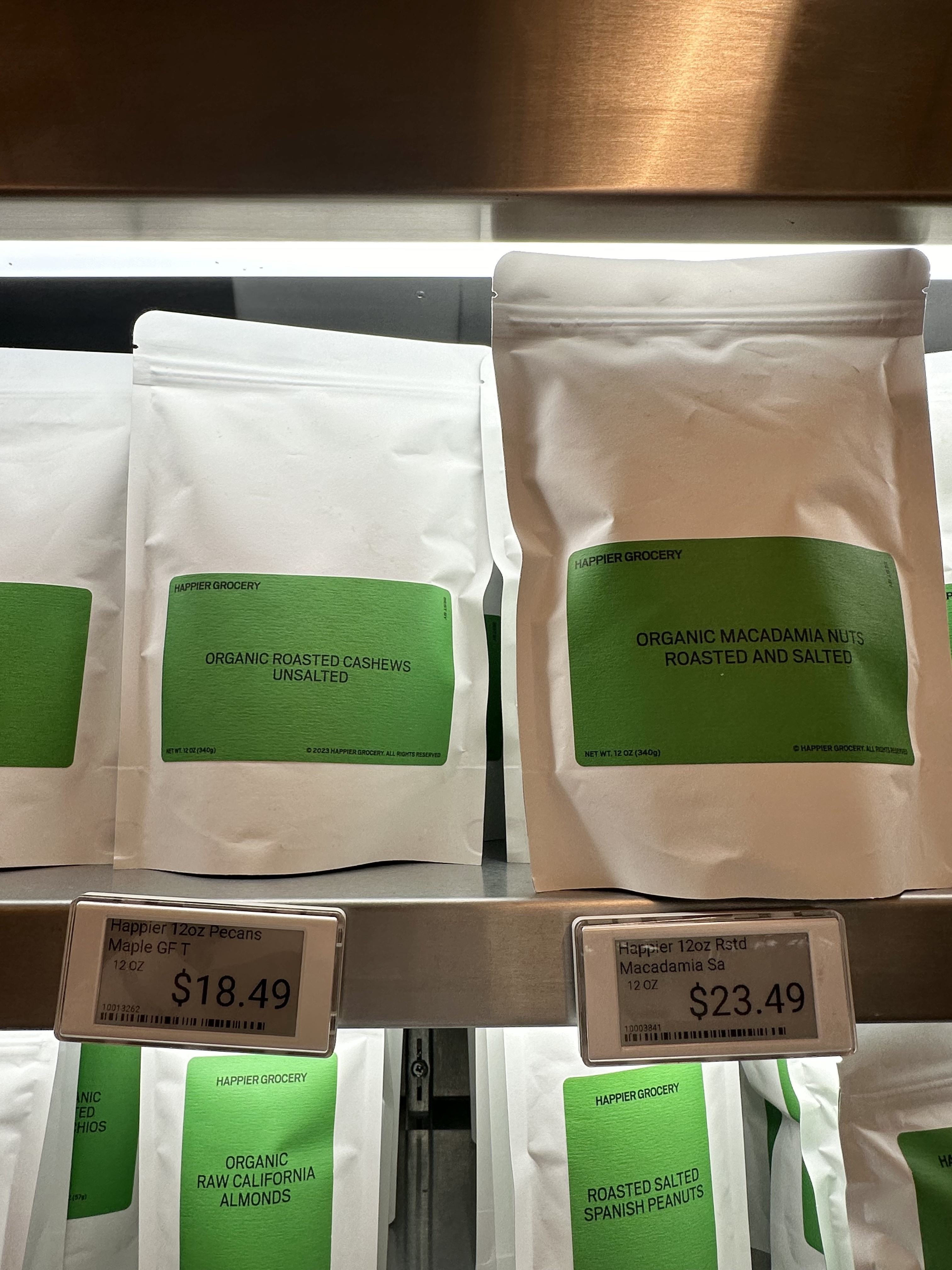 Organic roasted unsalted cashews and roasted and salted macadamia nuts in white bags with green labels on a store shelf with price tags