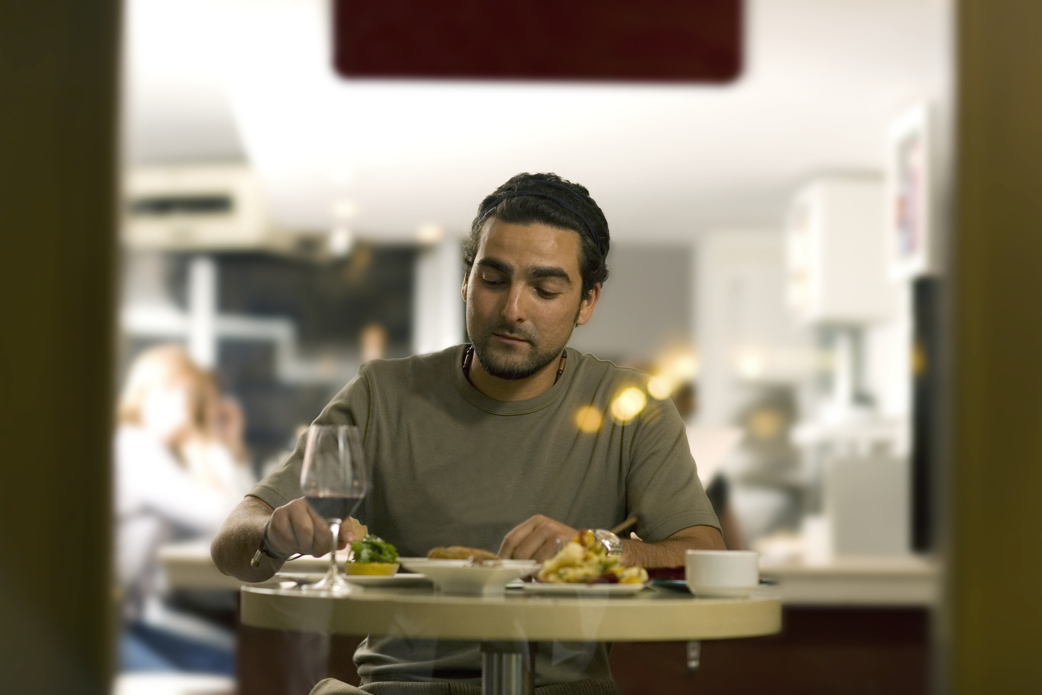 A man is seated at a table, dining alone in a restaurant with a plate of food and a drink in front of him. The restaurant appears busy in the background