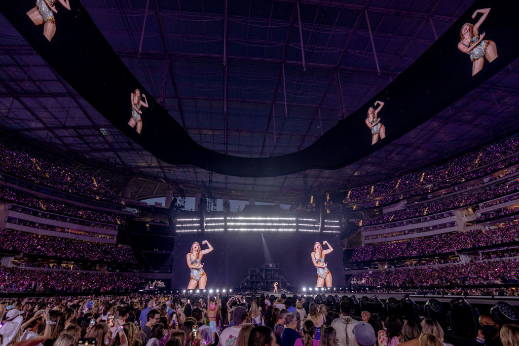Taylor Swift performs on stage at a stadium concert filled with fans, with large screens displaying her performance